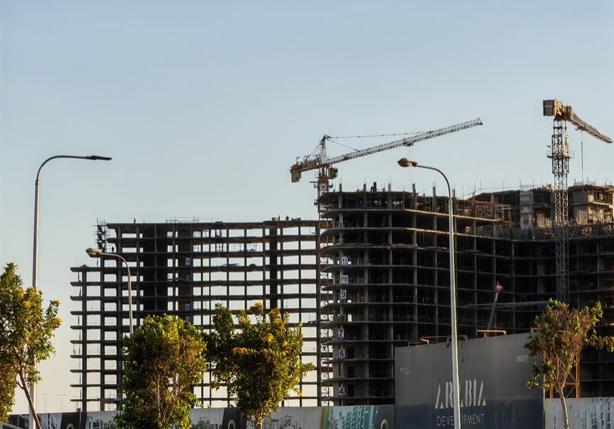 High-rise building under construction with cranes and trees in the forefront.